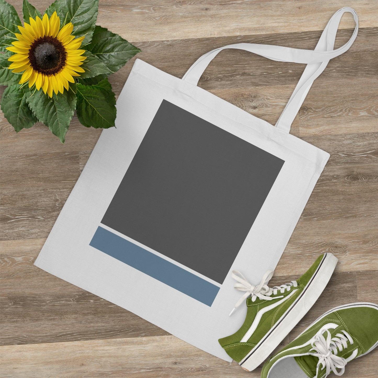 White tote bag with a dark gray square and blue stripe on a wooden surface with a sunflower and green shoes.