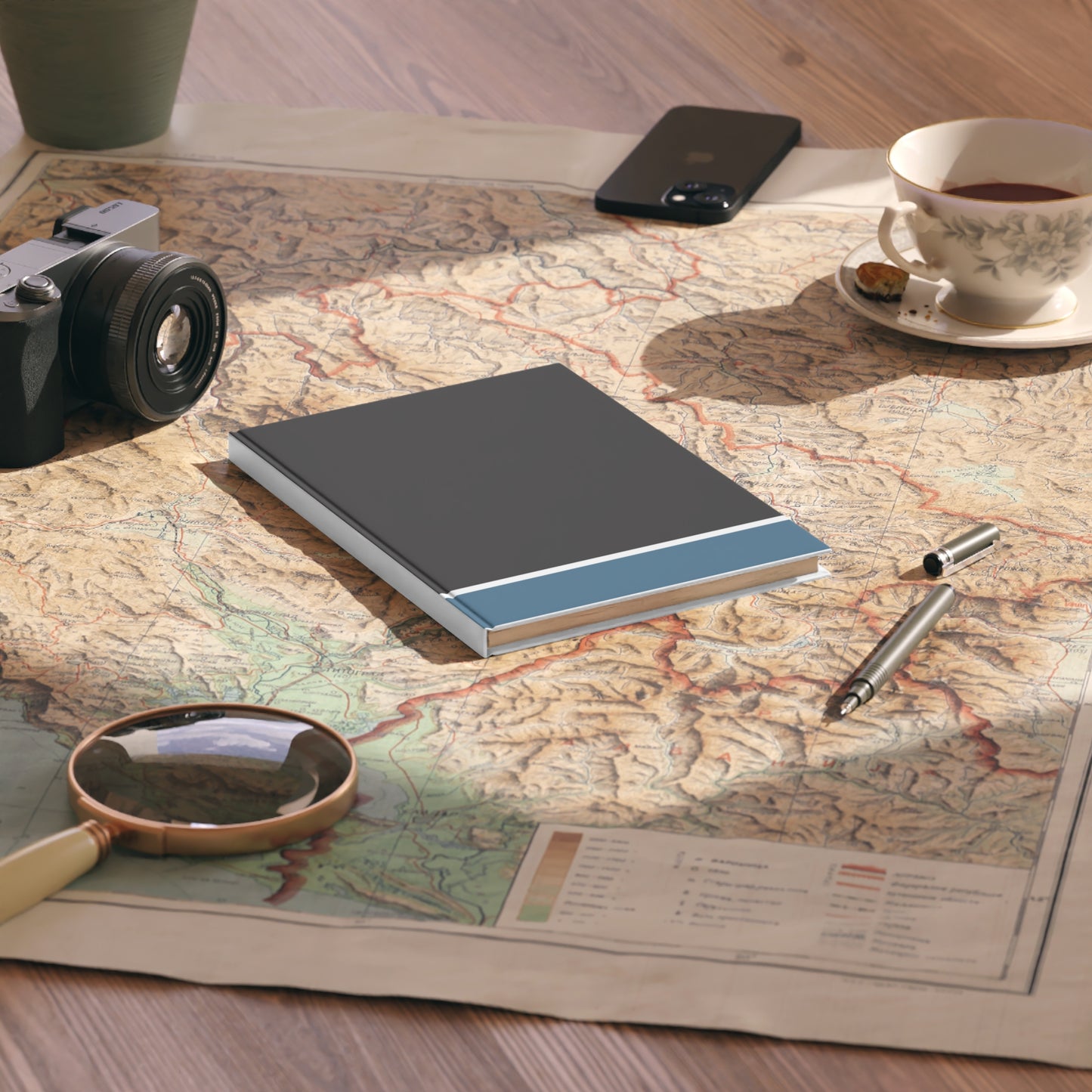 Notebook on a map with a camera, phone, cup of tea, and magnifying glass.