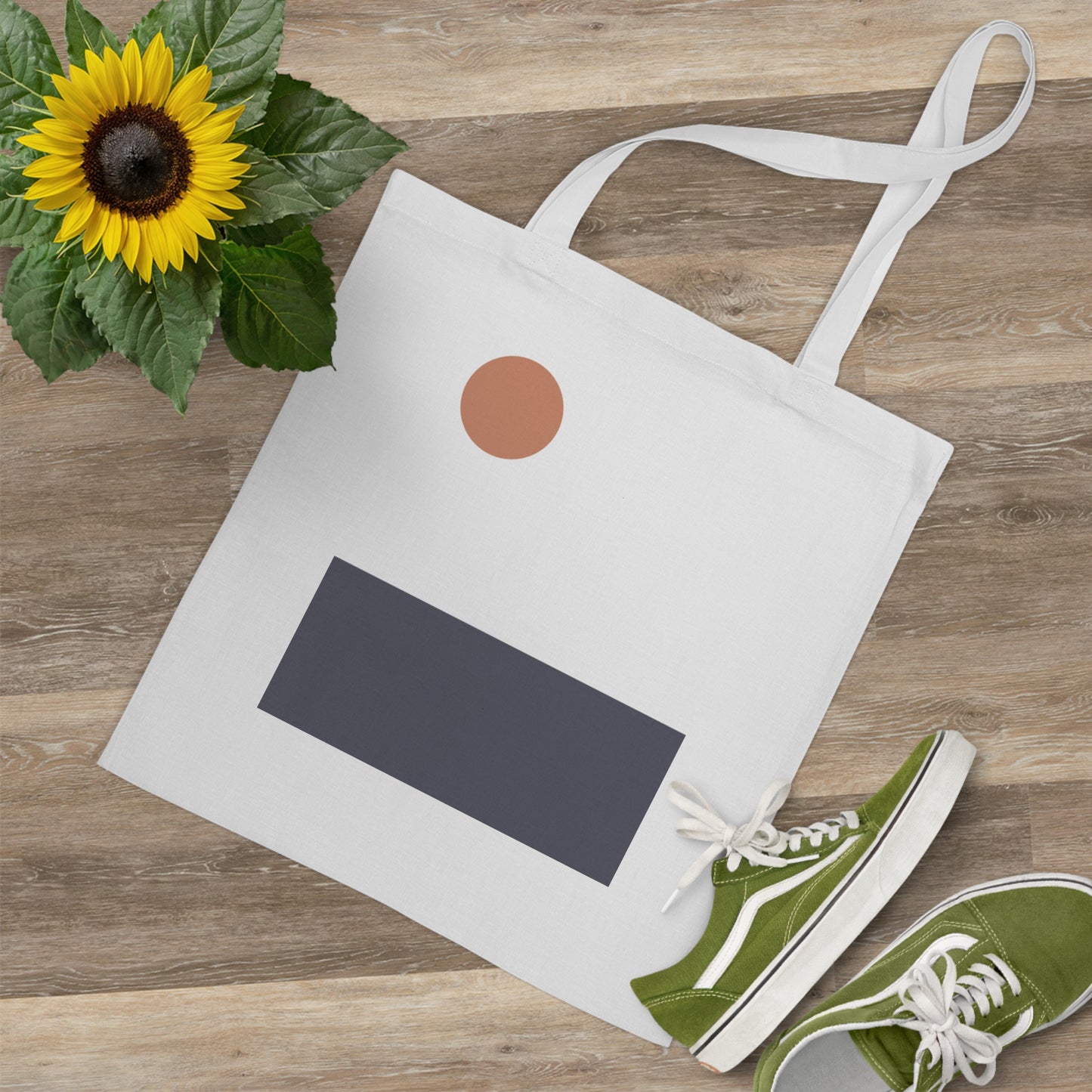 White tote bag with black label, green sneakers, sunflower, and brown circle on a wooden surface