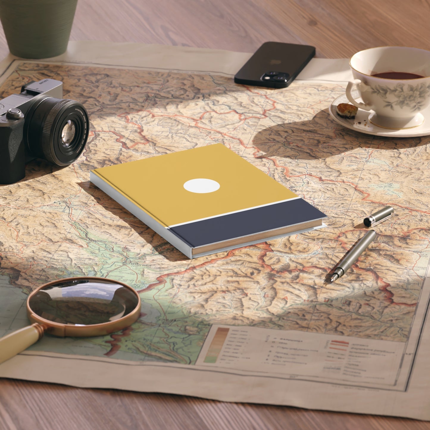 Notebook on a map with a camera, phone, cup of tea, pen, and magnifying glass.