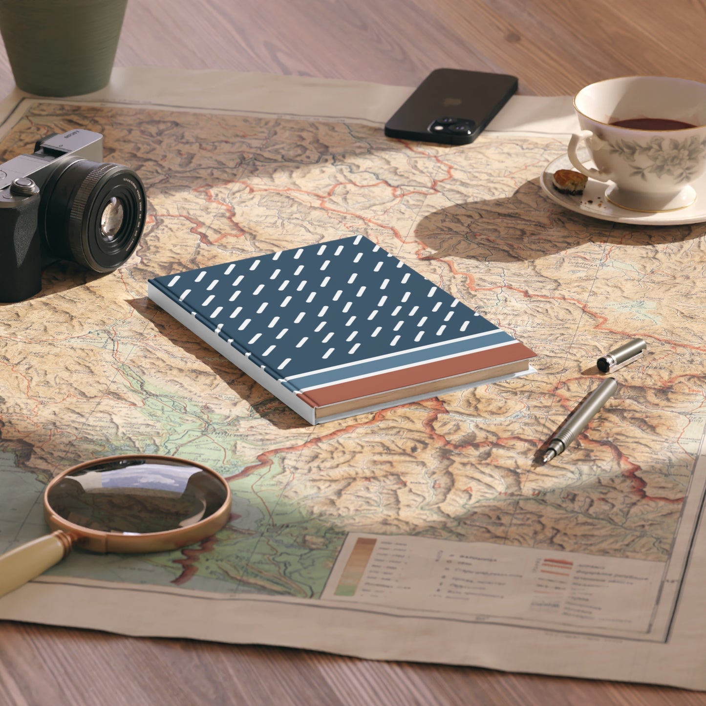 Notebook with a blue and red design on a map with a camera, phone, and tea cup nearby.
