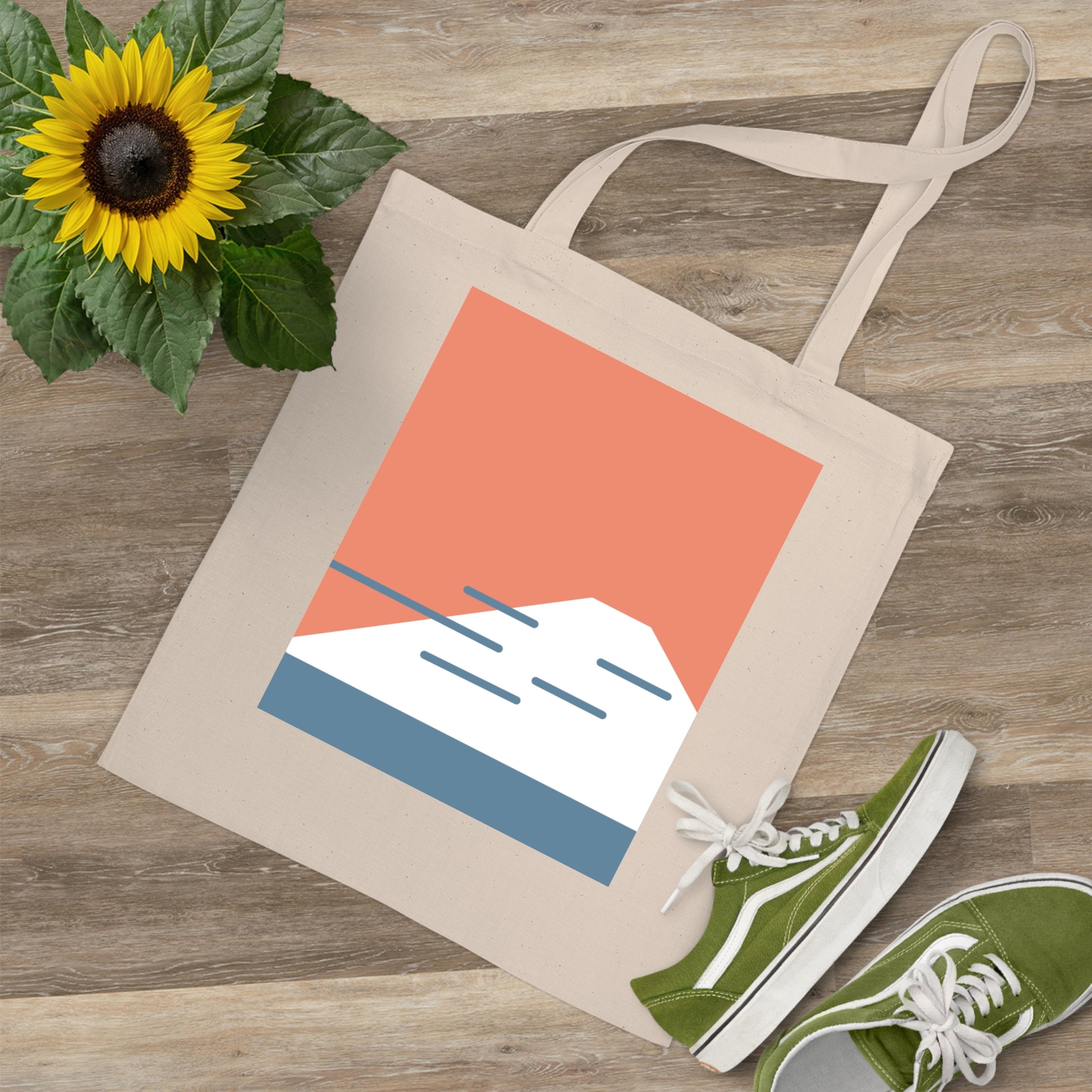 Beige tote bag with mountain design, green sneakers, and sunflower on wooden surface