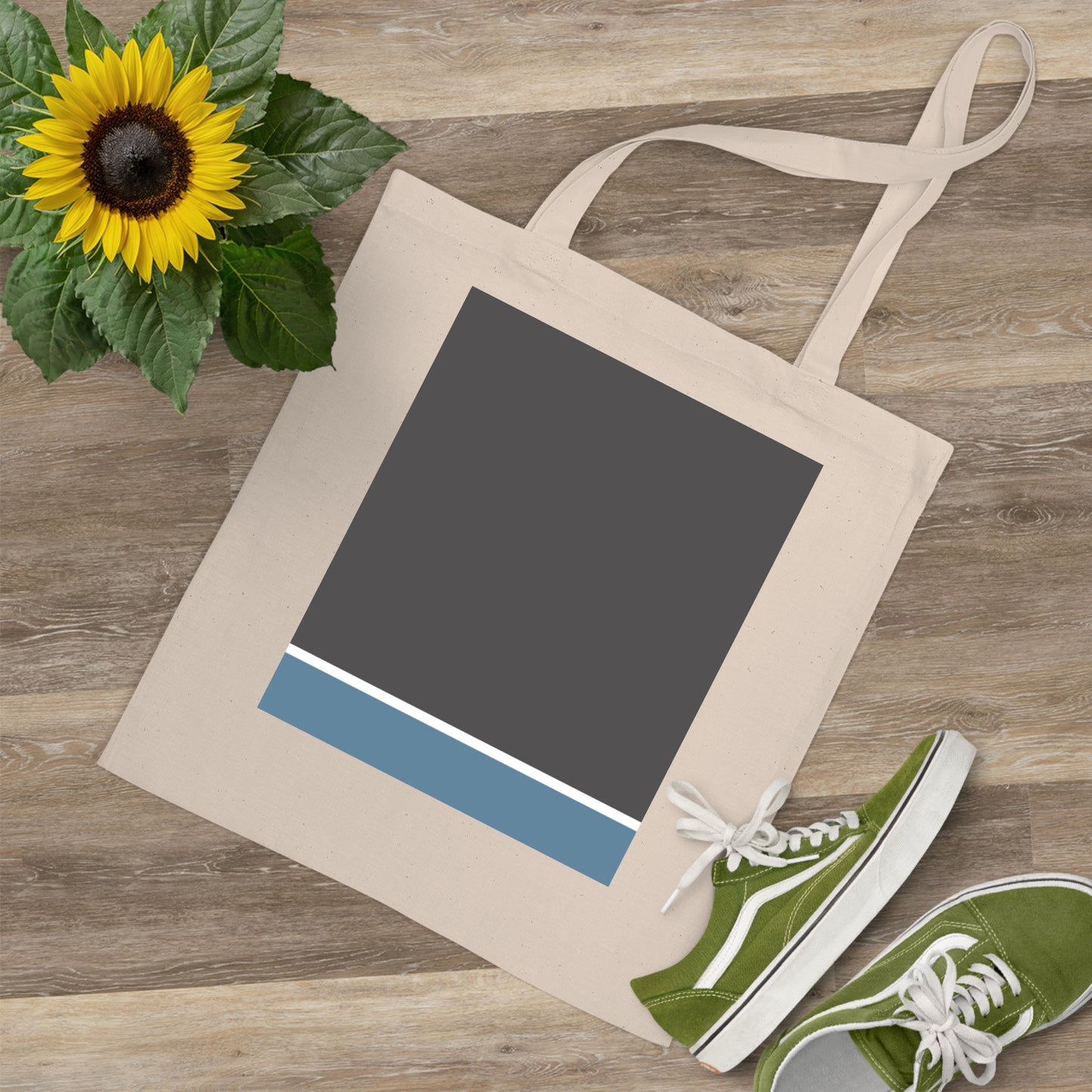 Beige tote bag with a dark gray panel and blue stripe, green sneakers, and sunflower on a wooden surface.