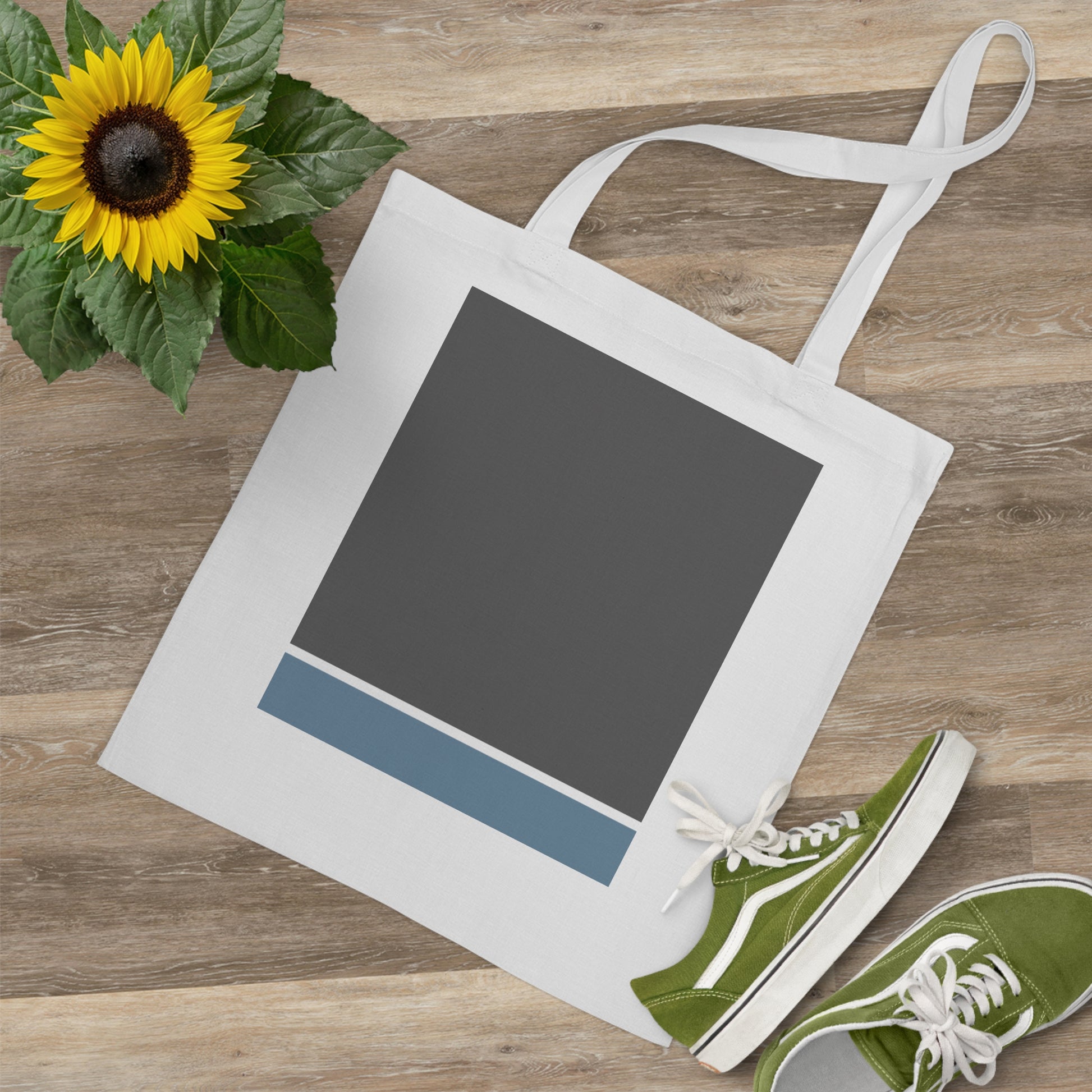 White tote bag with a dark gray square and blue stripe on a wooden surface with a sunflower and green shoes.