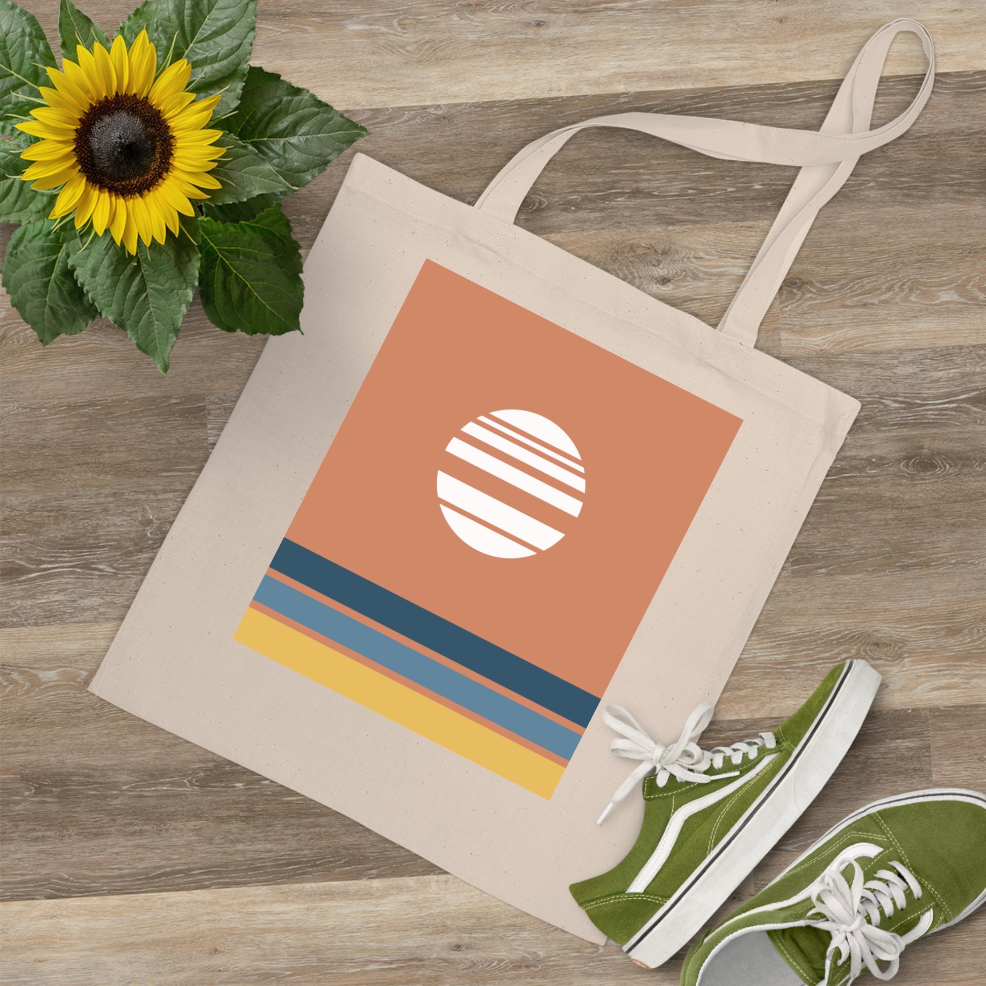 Beige tote bag with a colorful design on a wooden surface with green sneakers and a sunflower.